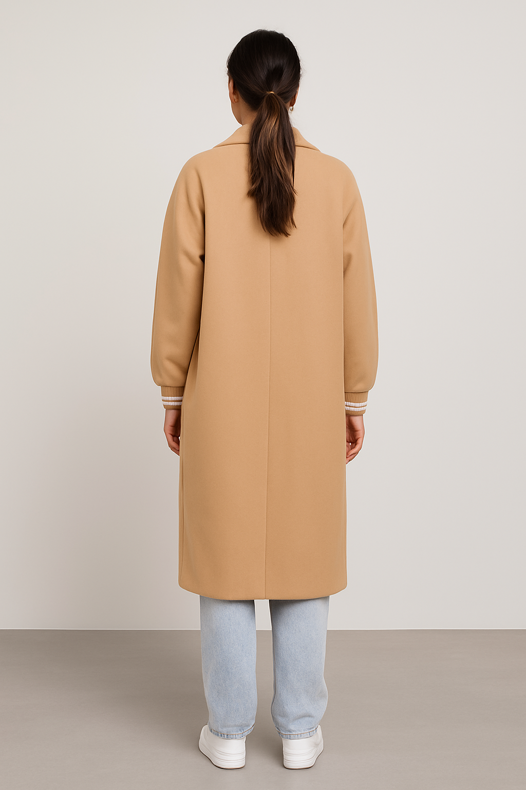 Back view of a woman wearing a beige ribbed cuff sleeves coat with light blue jeans and white sneakers against a plain background