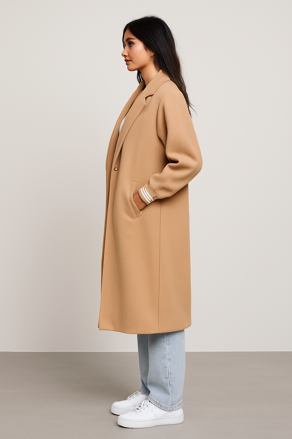 Side view of a woman wearing a beige ribbed cuff sleeves coat paired with light blue jeans and white sneakers, showcasing the coat's relaxed fit and elegant design