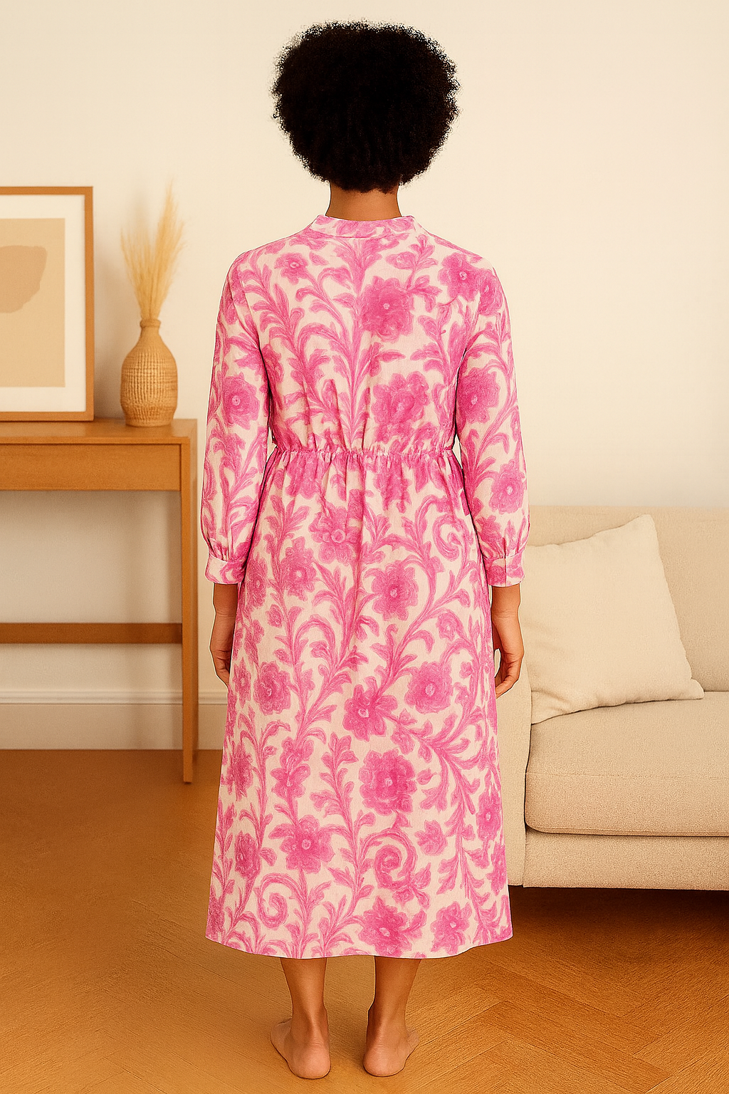 Back view of a woman wearing a pink floral Italian maxi dress made from lightweight viscose, standing in a cozy living room.